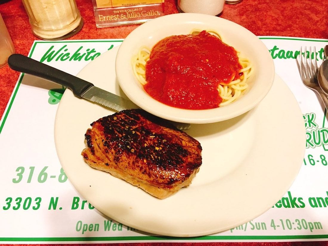 A plate with a grilled steak and a bowl of spaghetti topped with tomato sauce. A knife rests beside the steak; utensils and condiments are visible in the background. The plate is on a restaurant placemat.