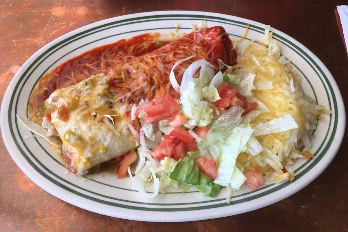 A plate with two enchiladas covered in red and green sauces and melted cheese, served with shredded lettuce, diced tomatoes, and more cheese on the side.
