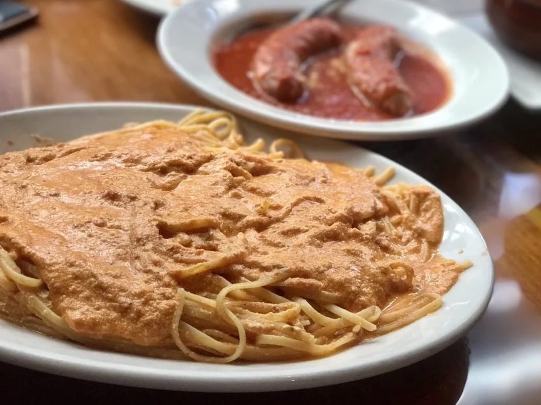 A plate of spaghetti topped with a creamy, orange-colored sauce sits in the foreground, while two sausages in tomato sauce are on a separate plate in the background.