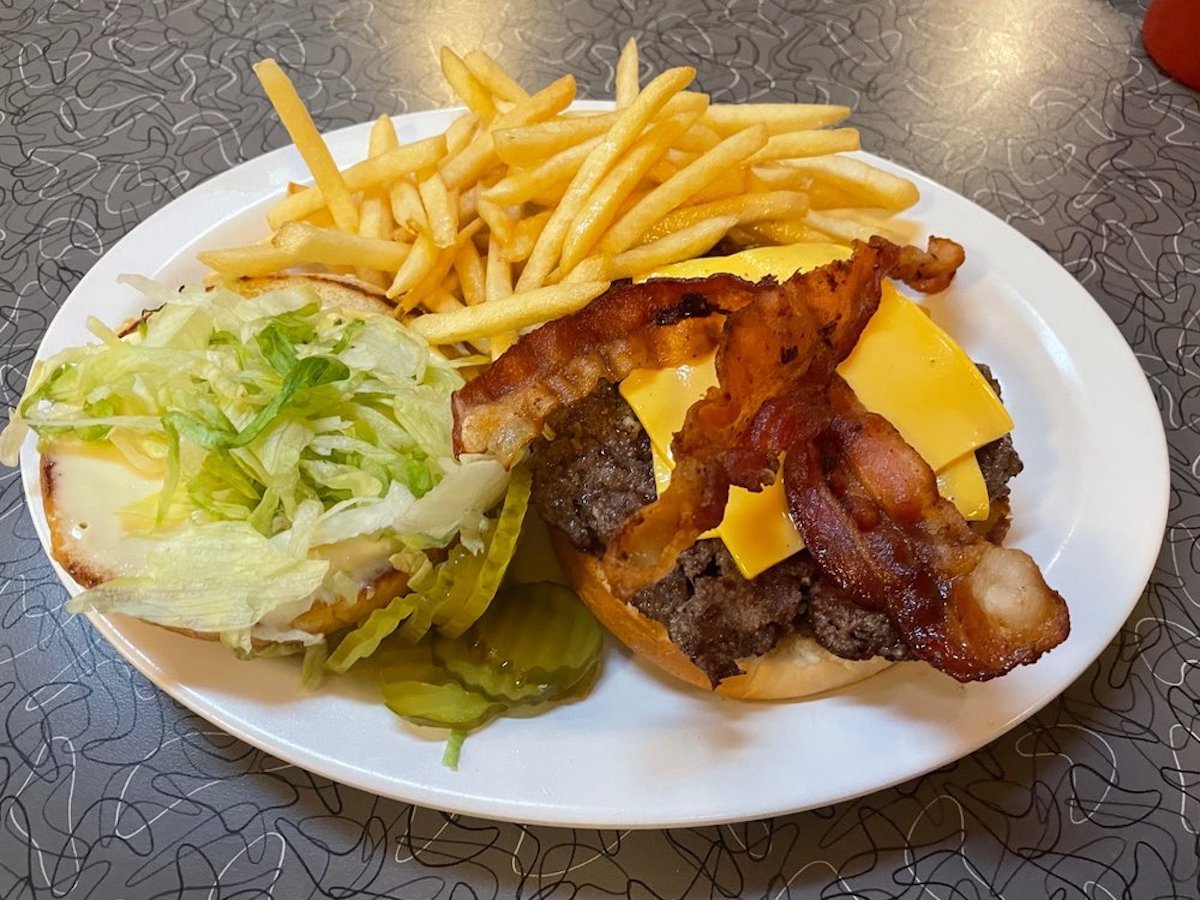 A plate with a cheeseburger topped with bacon, served with lettuce, pickles, and a side of French fries on a retro-patterned table.