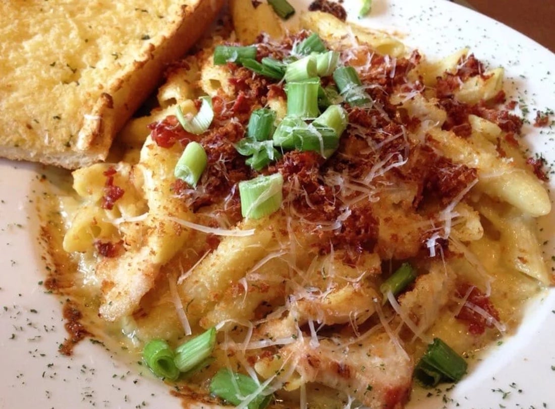 A plate of creamy penne pasta topped with grated cheese, bacon bits, and chopped green onions, served with a slice of garlic bread on the side.