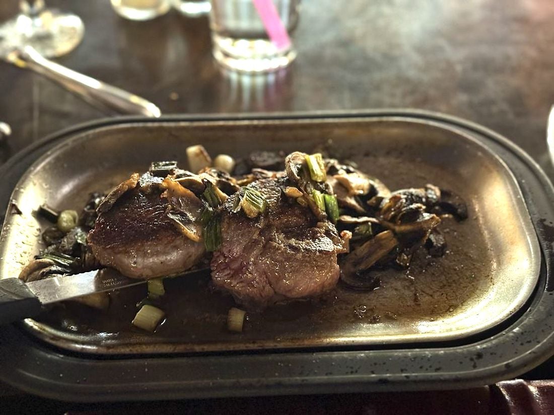 A cooked steak topped with sautéed mushrooms and green onions sits on a metal tray, echoing the quality of the best Italian restaurant near me. A glass of water and utensils rest on the dark table in the background.