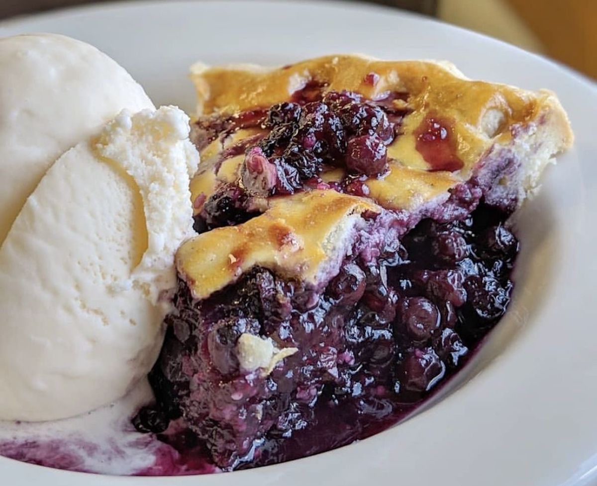 A slice of blueberry pie with a golden crust sits on a white plate, served with a scoop of vanilla ice cream melting at the side.