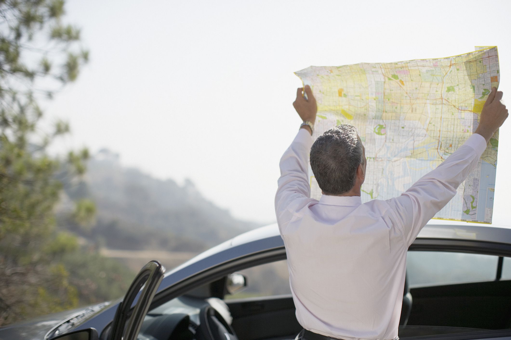 Man holding up road map with both arms stretched while reading it, standing against car with driver door opened with a blurred background of nature scenery