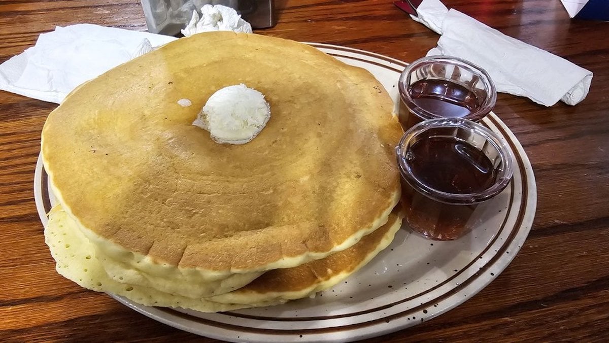 A stack of large, fluffy pancakes topped with a scoop of butter, served on a plate with two small cups of syrup on the side, placed on a wooden table with napkins in the background.