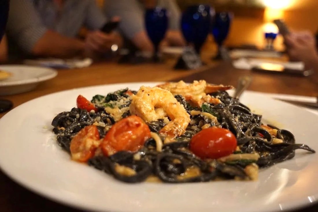 A plate of black squid ink pasta topped with shrimp, cherry tomatoes, and sauce sits on a table, with people blurred in the background holding phones.
