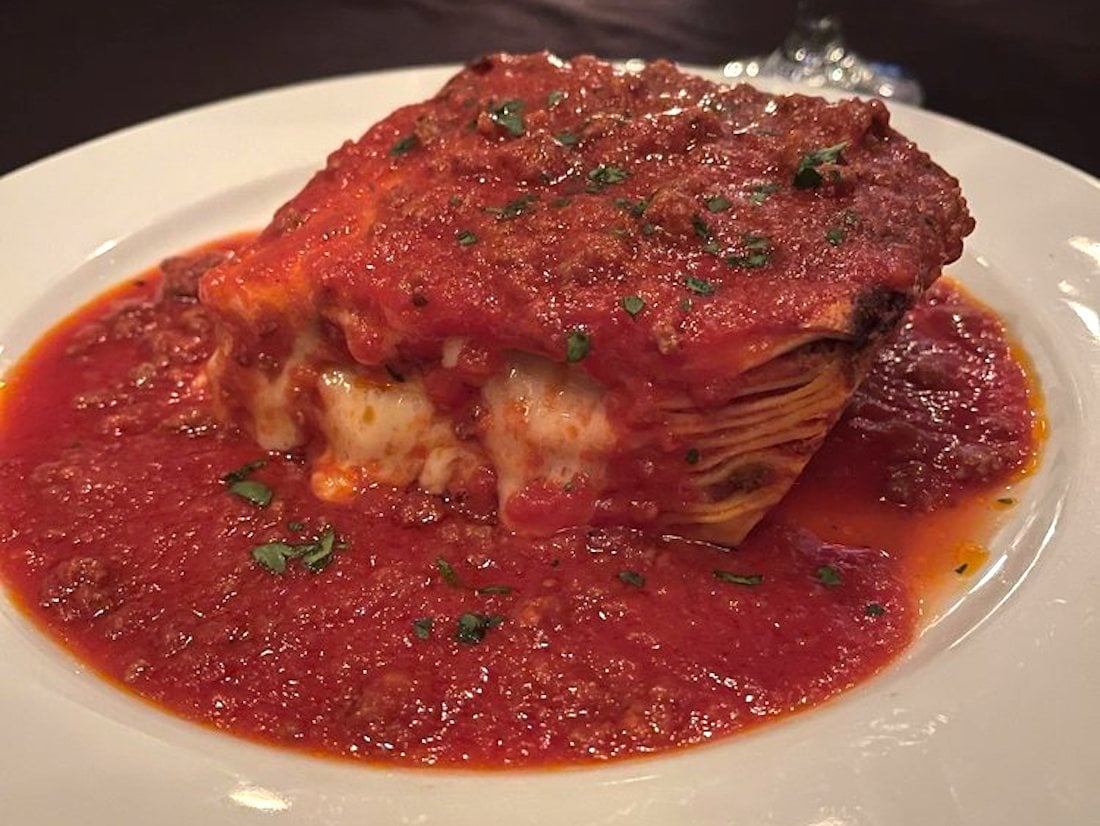 A slice of lasagna topped with rich tomato meat sauce, garnished with chopped herbs, sits on a white plate with extra sauce pooling around it. Cheese and layers of pasta are visible in the cut section.