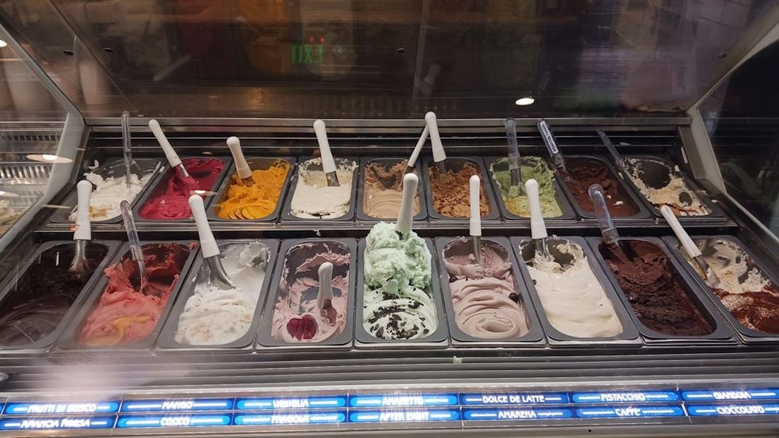 A display case filled with various flavors of gelato in metal trays, each with its own scoop. The gelato colors range from white and green to pink, brown, and yellow, with flavor labels visible below the trays.