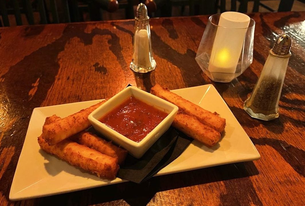 A rectangular plate with five mozzarella sticks arranged around a square dish of marinara sauce sits on a wooden table. Salt, pepper, and a candle are in the background.