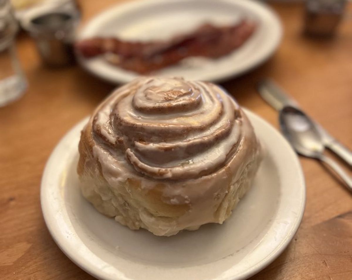 A close-up of a glazed cinnamon roll on a white plate, with another plate holding strips of bacon blurred in the background. A spoon and fork are visible on a wooden table.