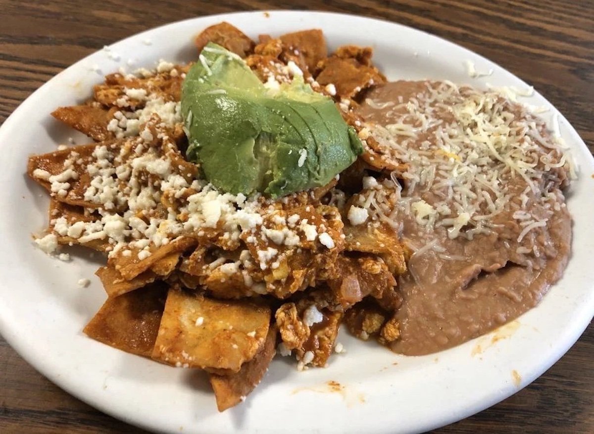 A plate of chilaquiles topped with crumbled cheese and sliced avocado, served with refried beans covered in shredded cheese on the side. The food is on a white plate on a wooden table.