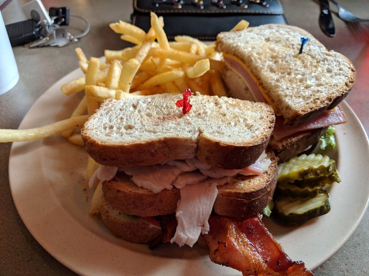 A plate with two deli sandwiches on whole wheat bread, each secured with a toothpick, served with French fries and pickle slices on the side. A wallet and keys are in the background.