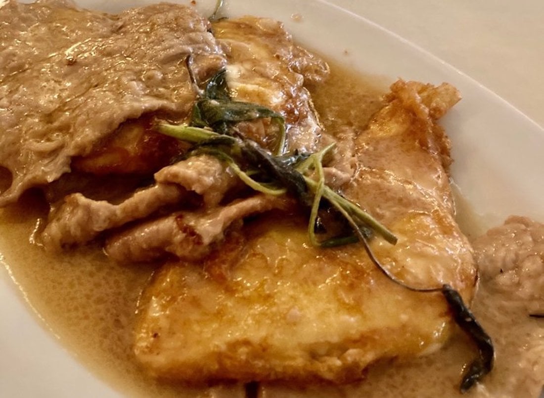 A close-up of a plate with slices of meat in a creamy brown sauce, garnished with cooked green herbs, next to golden-brown pieces of what appears to be pan-fried potatoes or fish.