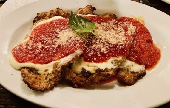 A plate of chicken parmesan topped with melted mozzarella cheese, marinara sauce, grated parmesan, and a basil leaf, served on a white dish.