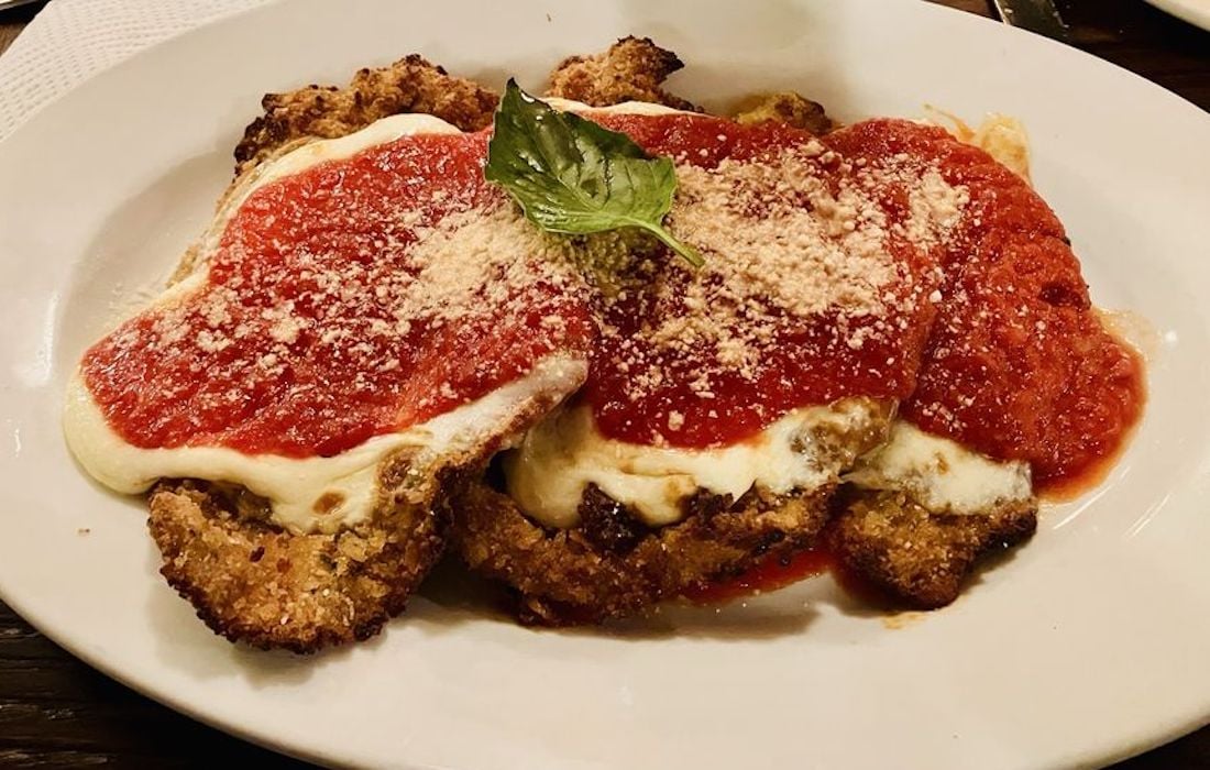 A plate of chicken parmesan topped with melted mozzarella cheese, marinara sauce, grated parmesan, and a basil leaf, served on a white dish.