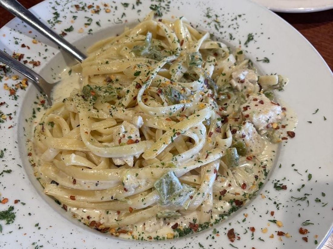A plate of fettuccine pasta in a creamy white sauce with herbs, red pepper flakes, and green vegetables, served with metal tongs on a white dish.