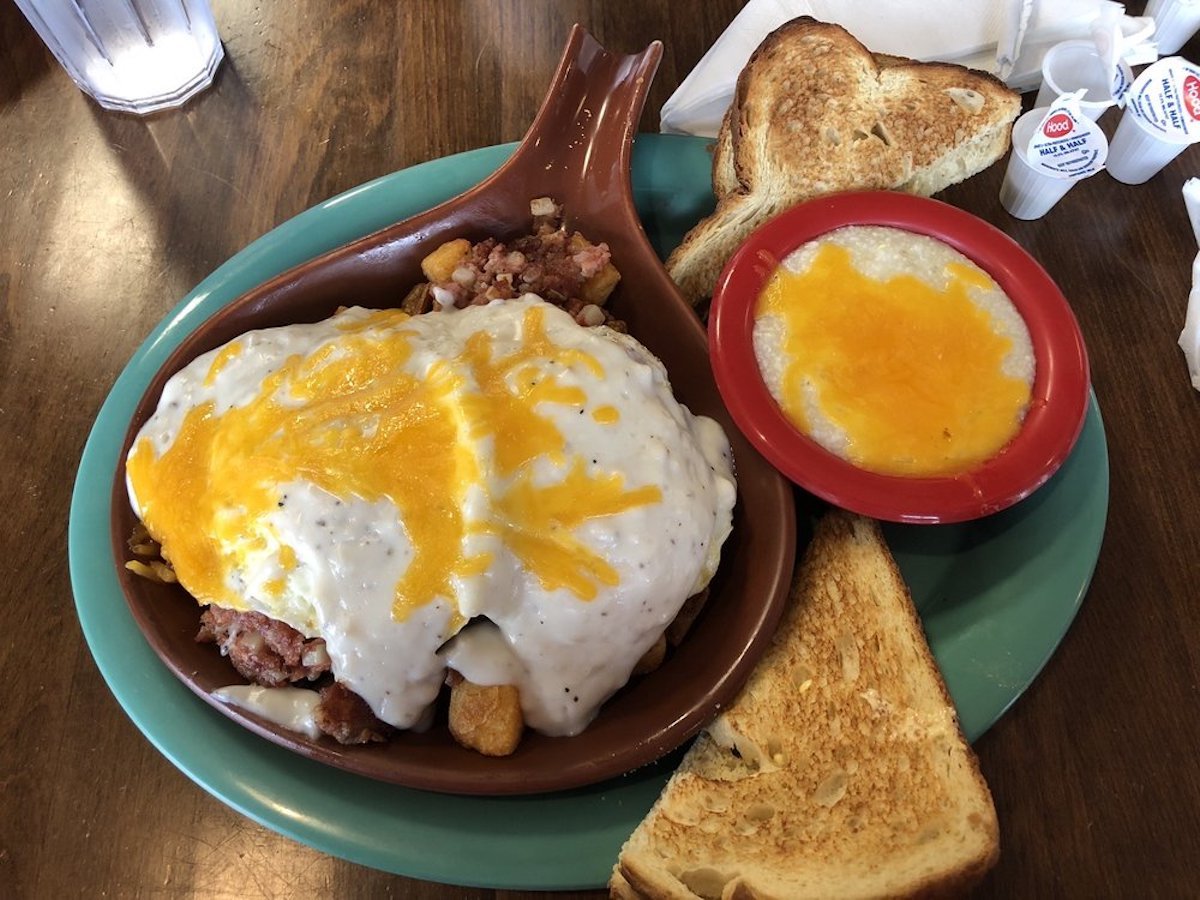 A plate with toast, a dish of cheesy grits, and a large portion of hash browns topped with creamy gravy and melted cheddar cheese on a wooden table. A glass of water and packets of jelly are in the background.