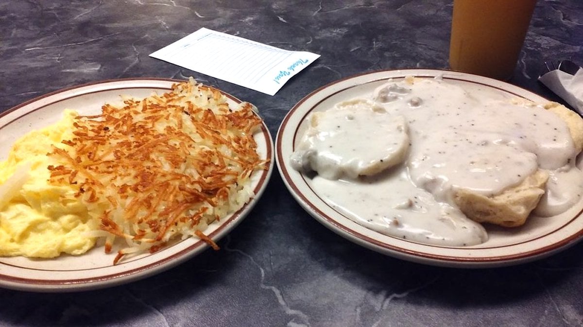 Two plates on a table: one with scrambled eggs and crispy hash browns, the other with biscuits covered in white sausage gravy. There is a cup and a receipt in the background.