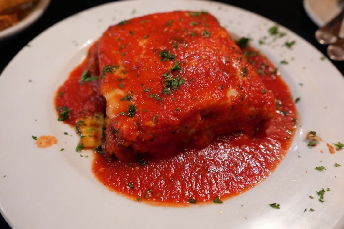 A slice of lasagna topped with melted cheese and tomato sauce, garnished with chopped parsley, served on a white plate.