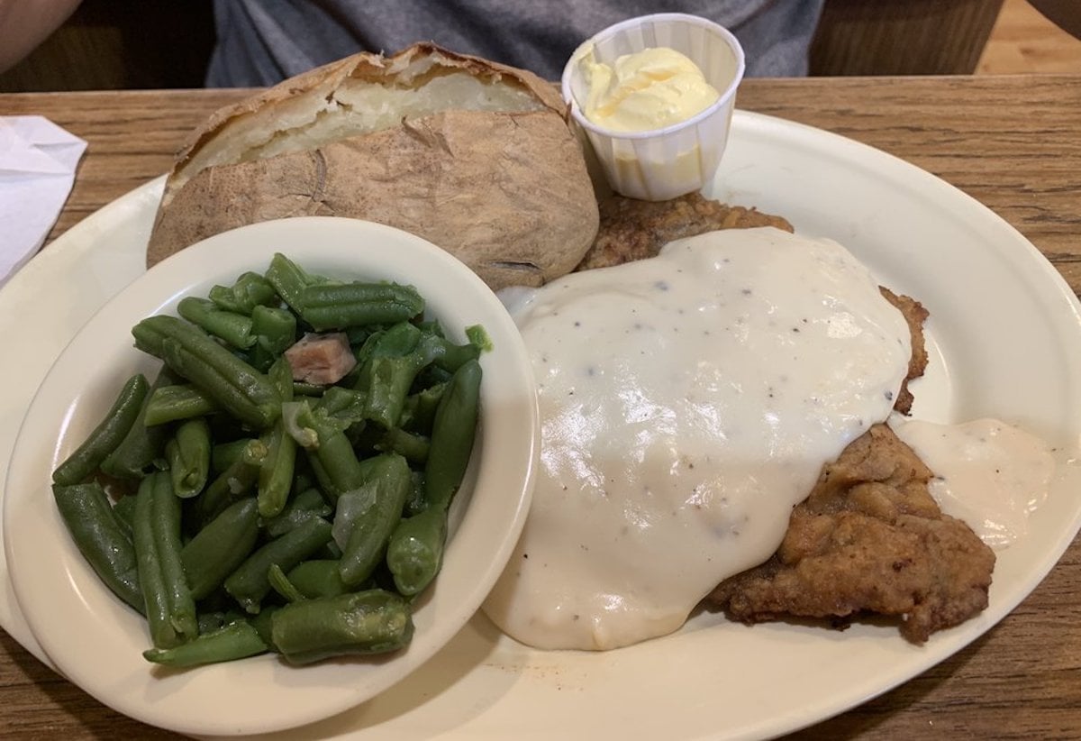 A plate with chicken fried steak covered in white gravy, a baked potato with a dollop of butter, and a side of green beans with bits of bacon, all on a wooden table.