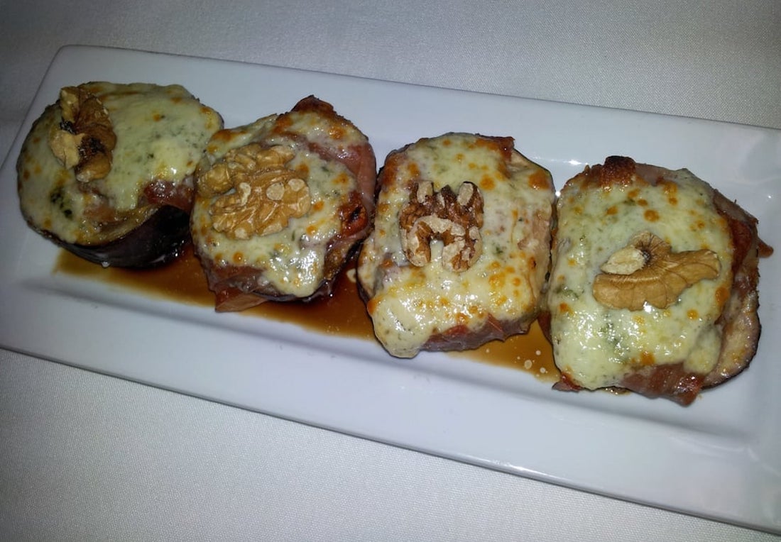 Four pieces of steak wrapped in prosciutto, topped with melted cheese and walnut halves, served on a rectangular white plate with a dark sauce underneath.