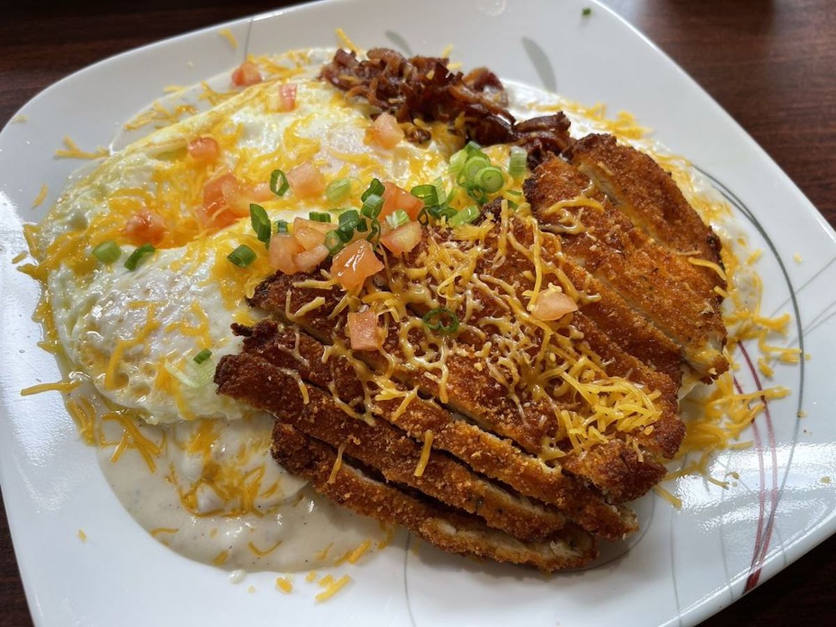 A plate of crispy breaded chicken cutlets topped with shredded cheddar cheese, diced tomatoes, and green onions, served over creamy white gravy with two fried eggs and a side of caramelized onions.