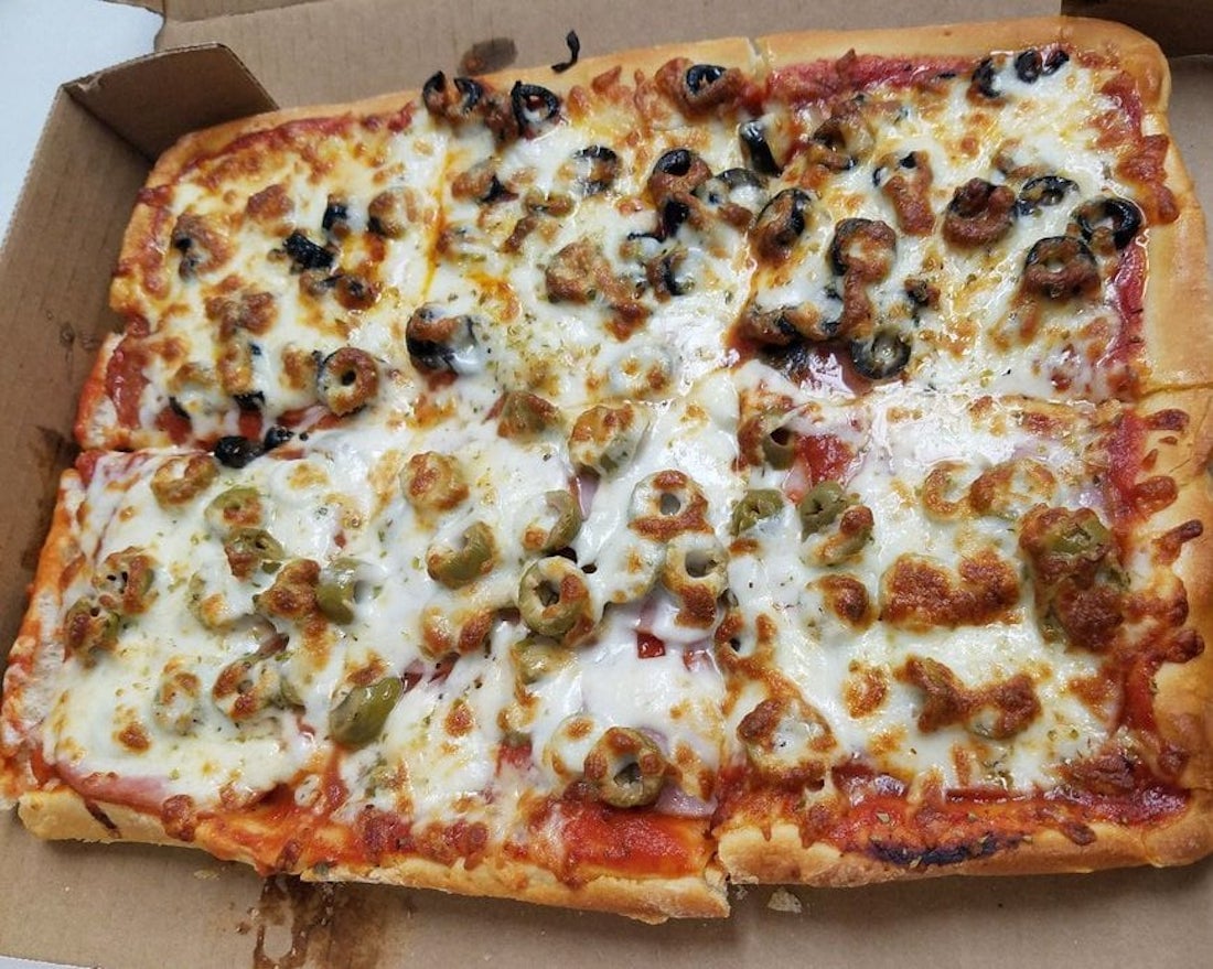 Rectangular pizza cut into eight square slices, topped with melted cheese, black olives, and green olives, in an open cardboard box. The pizza has a golden-brown crust and visible tomato sauce.