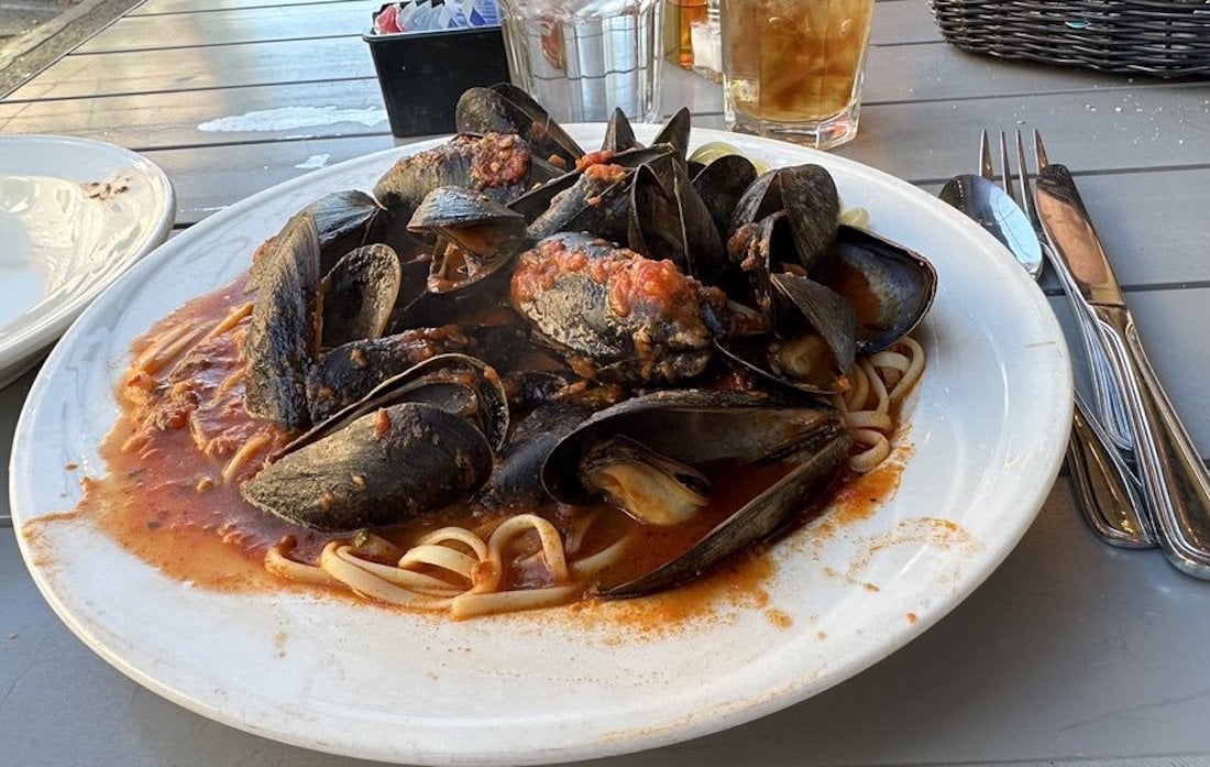 A white plate of linguine pasta topped with mussels in a red tomato-based sauce sits on an outdoor table, with utensils, a drink, and condiments visible in the background.