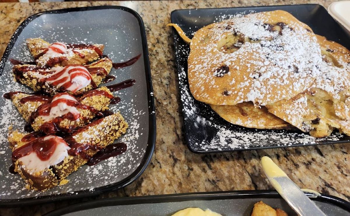 Two plates of food on a granite countertop: one with pancakes dusted with powdered sugar, the other with breaded French toast slices topped with whipped cream and drizzled with berry sauce.
