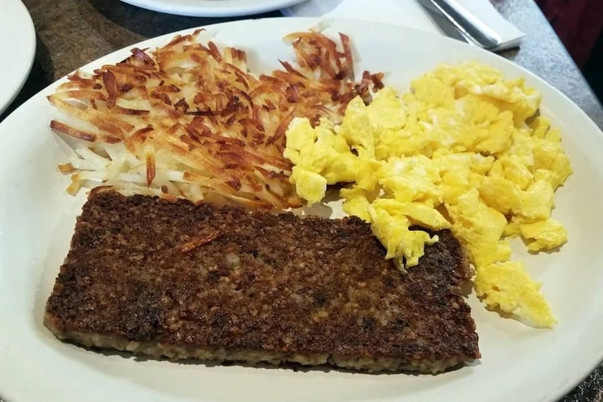 A breakfast plate with scrambled eggs, crispy hash browns, and a rectangular slice of browned sausage or meatloaf, served on a white dish.