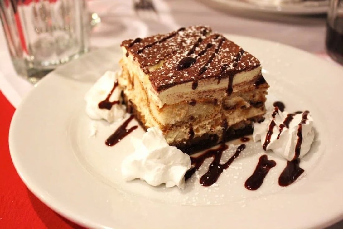 A slice of tiramisu topped with cocoa powder and powdered sugar, served on a white plate with whipped cream dollops and chocolate sauce drizzle.