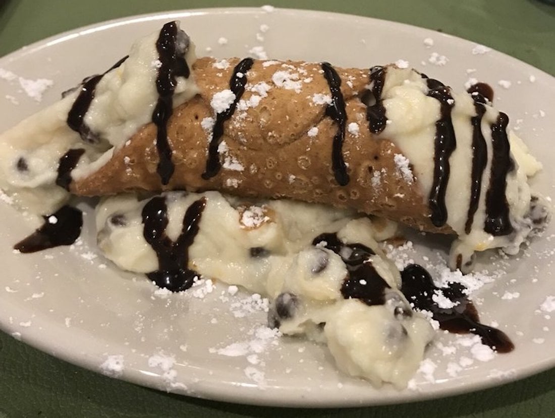 A cannoli with a crisp shell filled with creamy ricotta, chocolate chips, and topped with chocolate drizzle and powdered sugar, served on a white plate.
