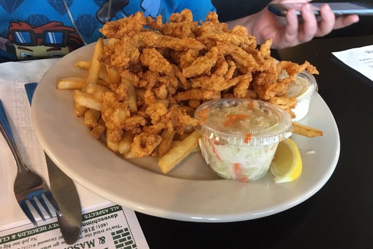 A plate piled high with fried clam strips on top of French fries, served with a portion of coleslaw in a plastic cup and a lemon wedge. A person’s hand holding a phone is visible in the background.
