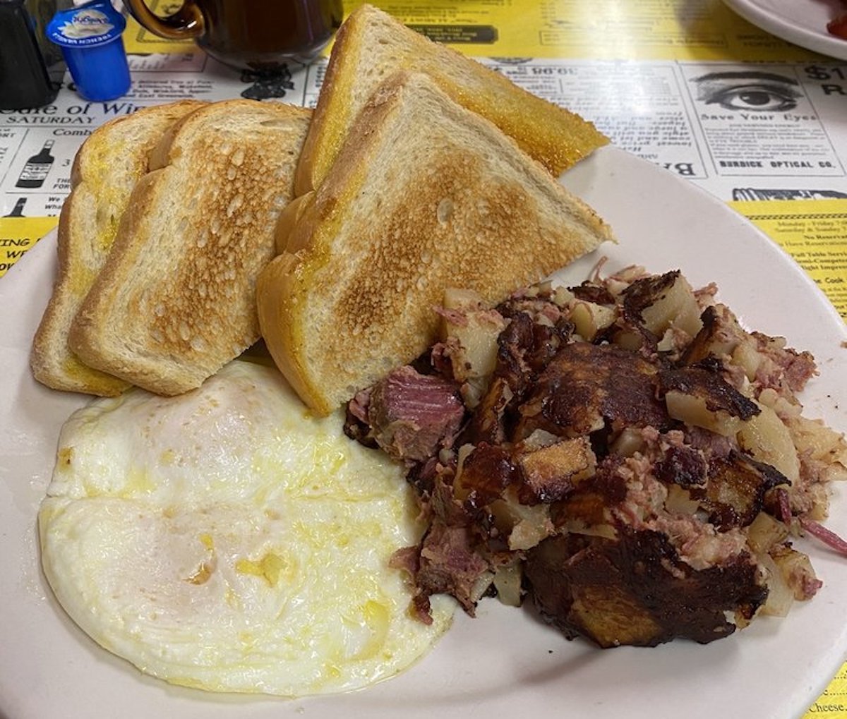 A plate with four slices of toasted bread, two fried eggs, and a serving of corned beef hash made with chopped beef and potatoes. The food is on a white plate atop a table with a yellow placemat.