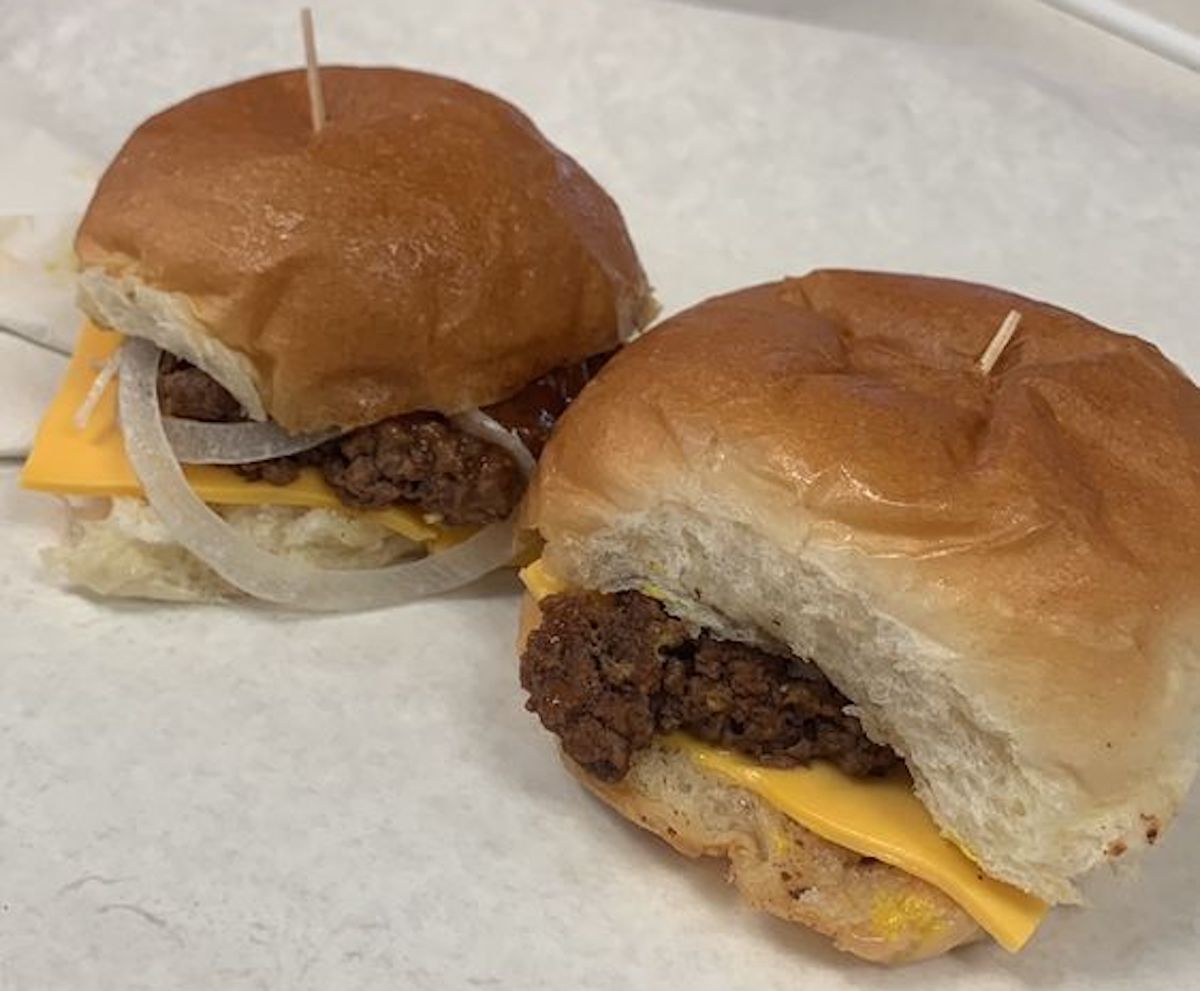 Two cheeseburgers on parchment paper, each with a beef patty, yellow cheese, and raw onion slices on a soft bun—one with a bite taken out—just like you'd find at the best hole-in-the-wall diners.