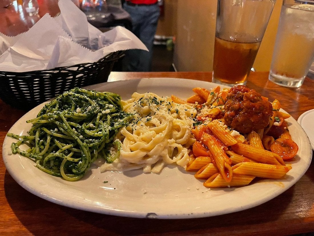 A plate with three kinds of pasta: pesto spaghetti, fettuccine alfredo, and penne with marinara sauce and meatballs, sits on a wooden table with drinks and a basket of bread in the background.
