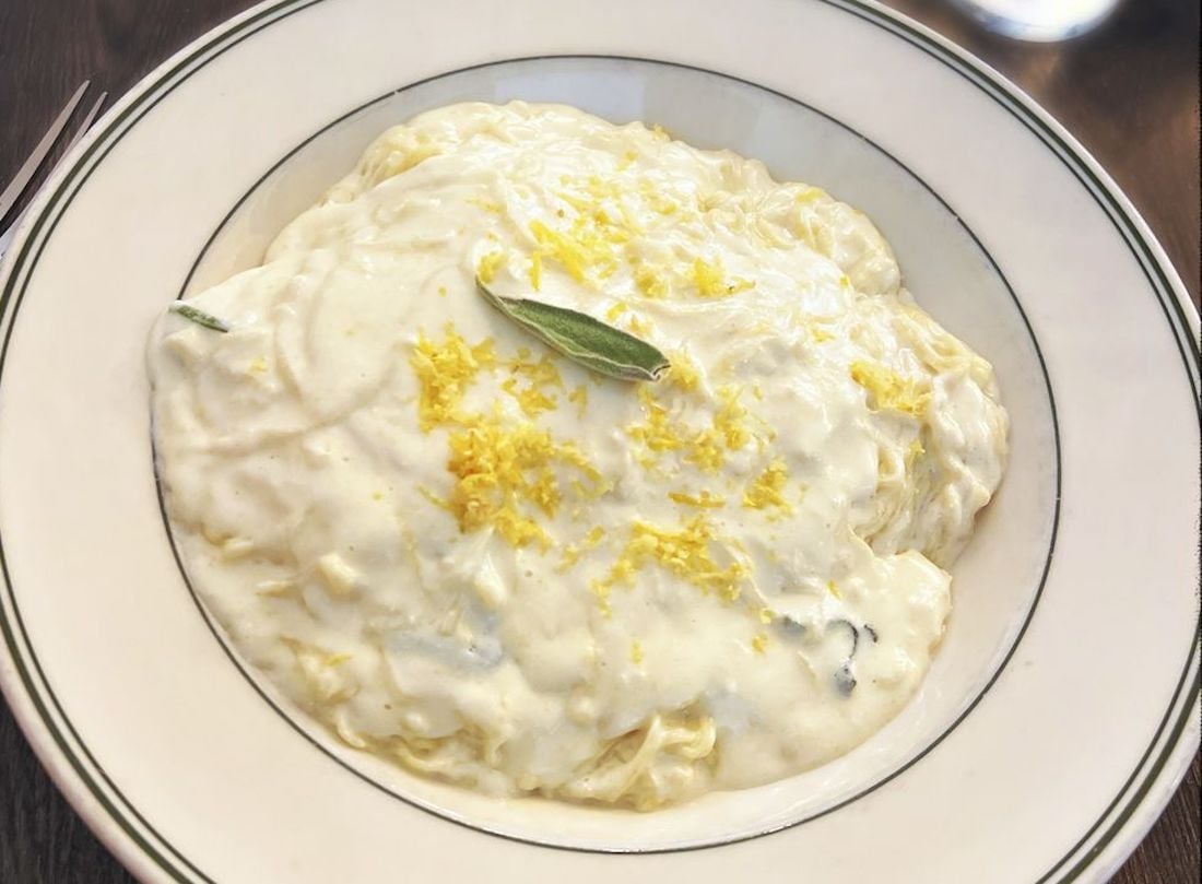A plate of pasta covered in creamy white sauce, garnished with lemon zest and a fresh sage leaf, served in a white bowl with a thin black rim.