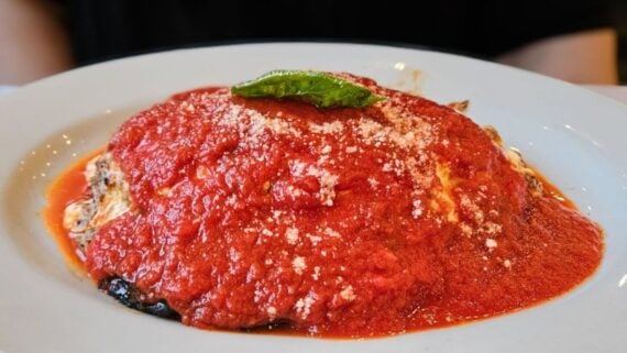 A plate of eggplant parmesan topped with chunky red tomato sauce, grated cheese, and a fresh basil leaf, served on a white dish against a dark background.