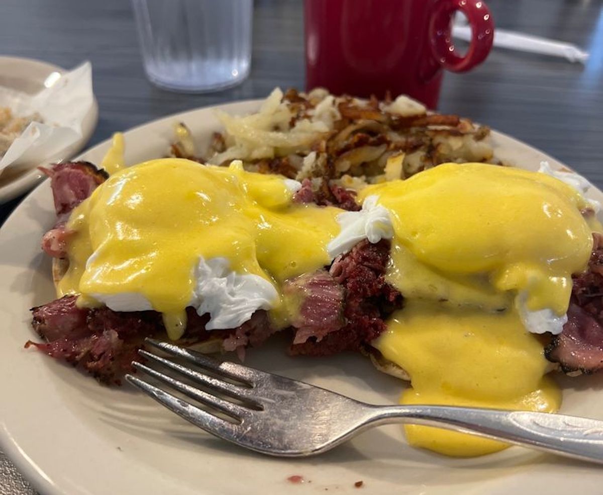 A plate of eggs Benedict with hollandaise sauce over poached eggs and corned beef on English muffins, served with hash browns—a classic breakfast you’d find at the best hole-in-the-wall diners. A fork rests on the plate; a red mug and glass of water are nearby.