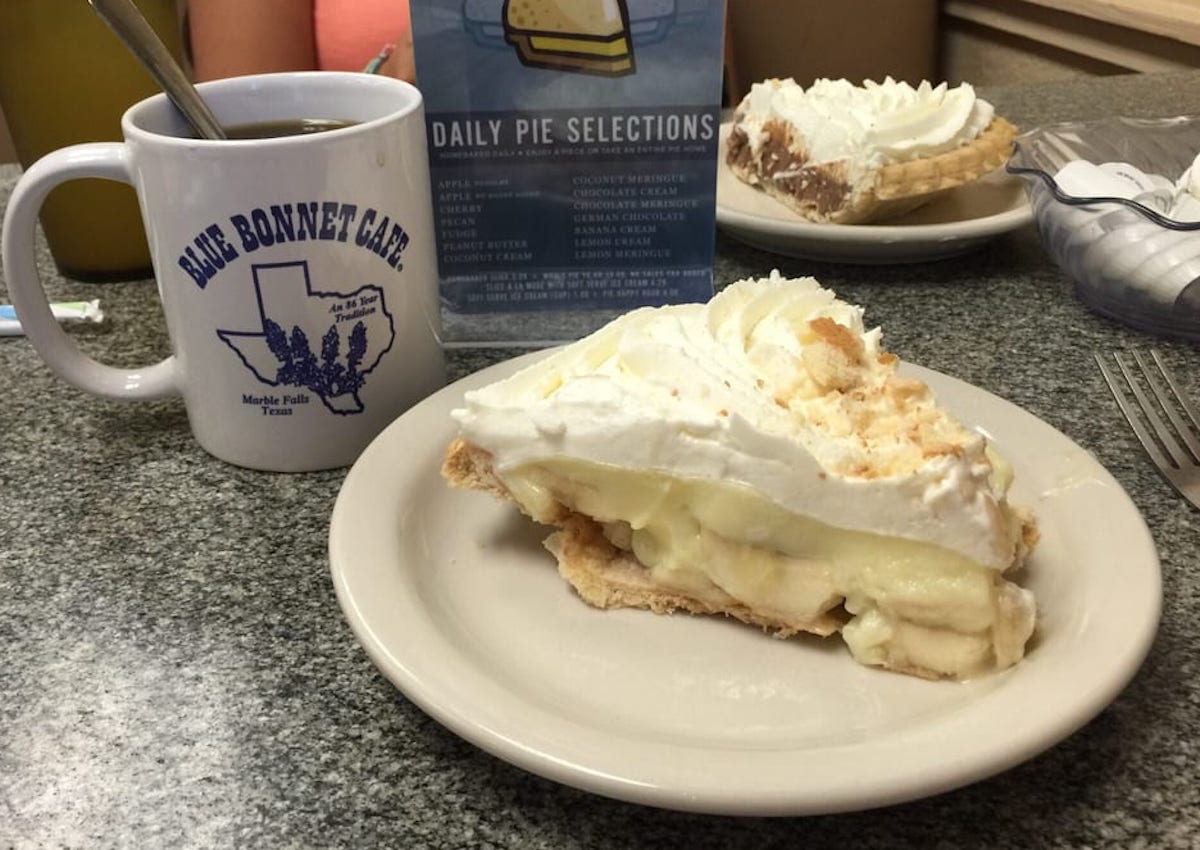 A slice of cream pie with a flaky crust and whipped topping sits on a plate next to a mug labeled "Blue Bonnet Cafe" and a menu displaying daily pie selections, all on a speckled tabletop.