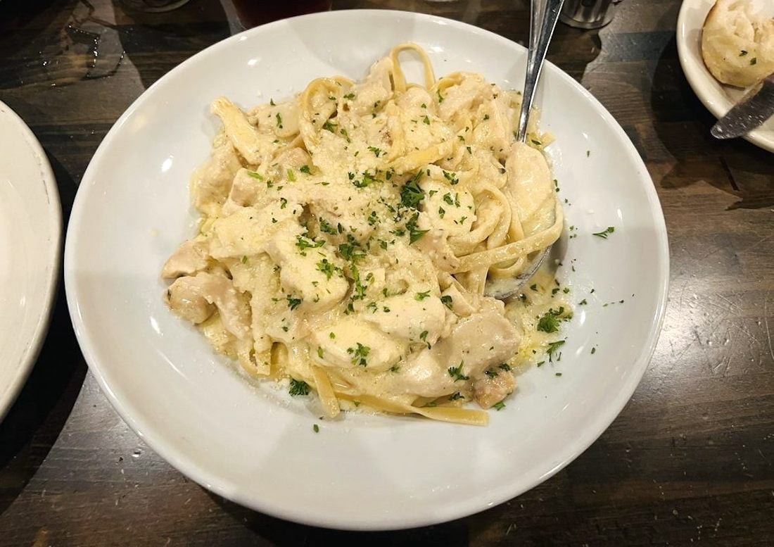 A white bowl filled with creamy fettuccine Alfredo pasta topped with chopped herbs, featuring pieces of chicken. A fork is placed in the bowl, and the dish is set on a dark wooden table.
