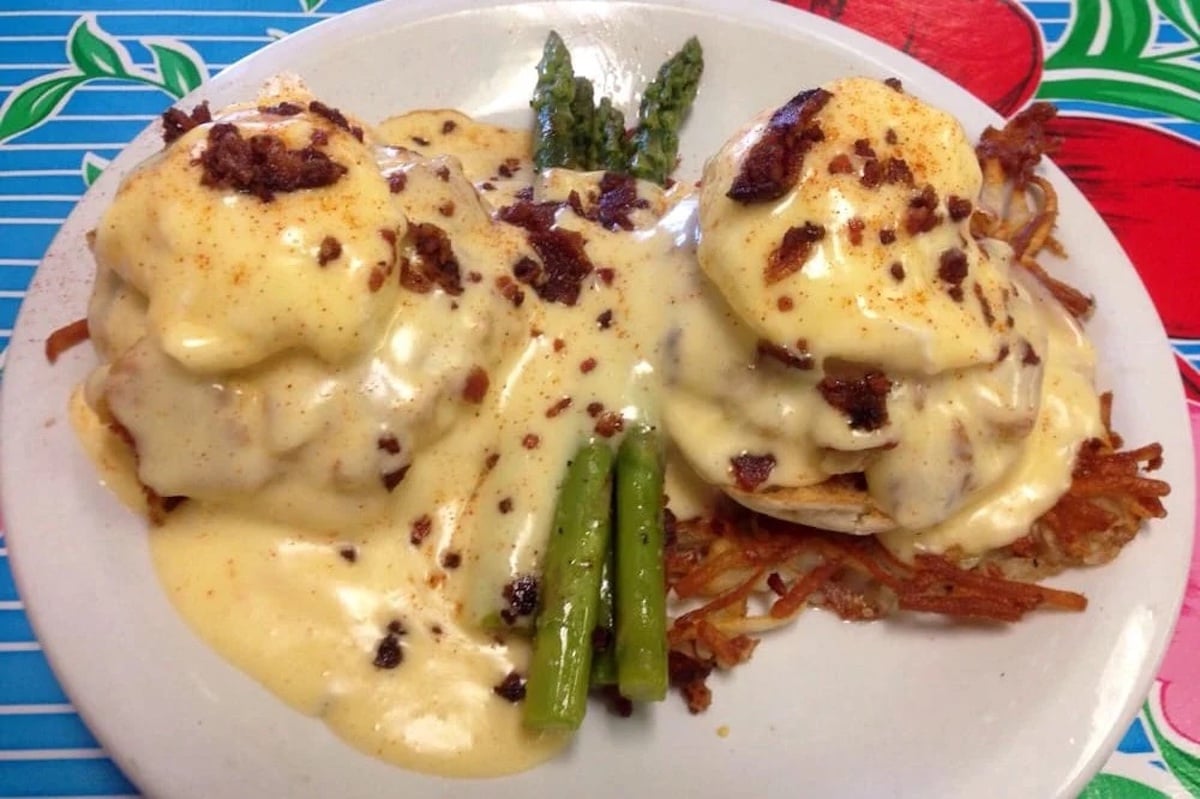 A plate of eggs Benedict topped with creamy hollandaise sauce and bacon bits, served on hash browns with a few asparagus spears, on a colorful tablecloth.