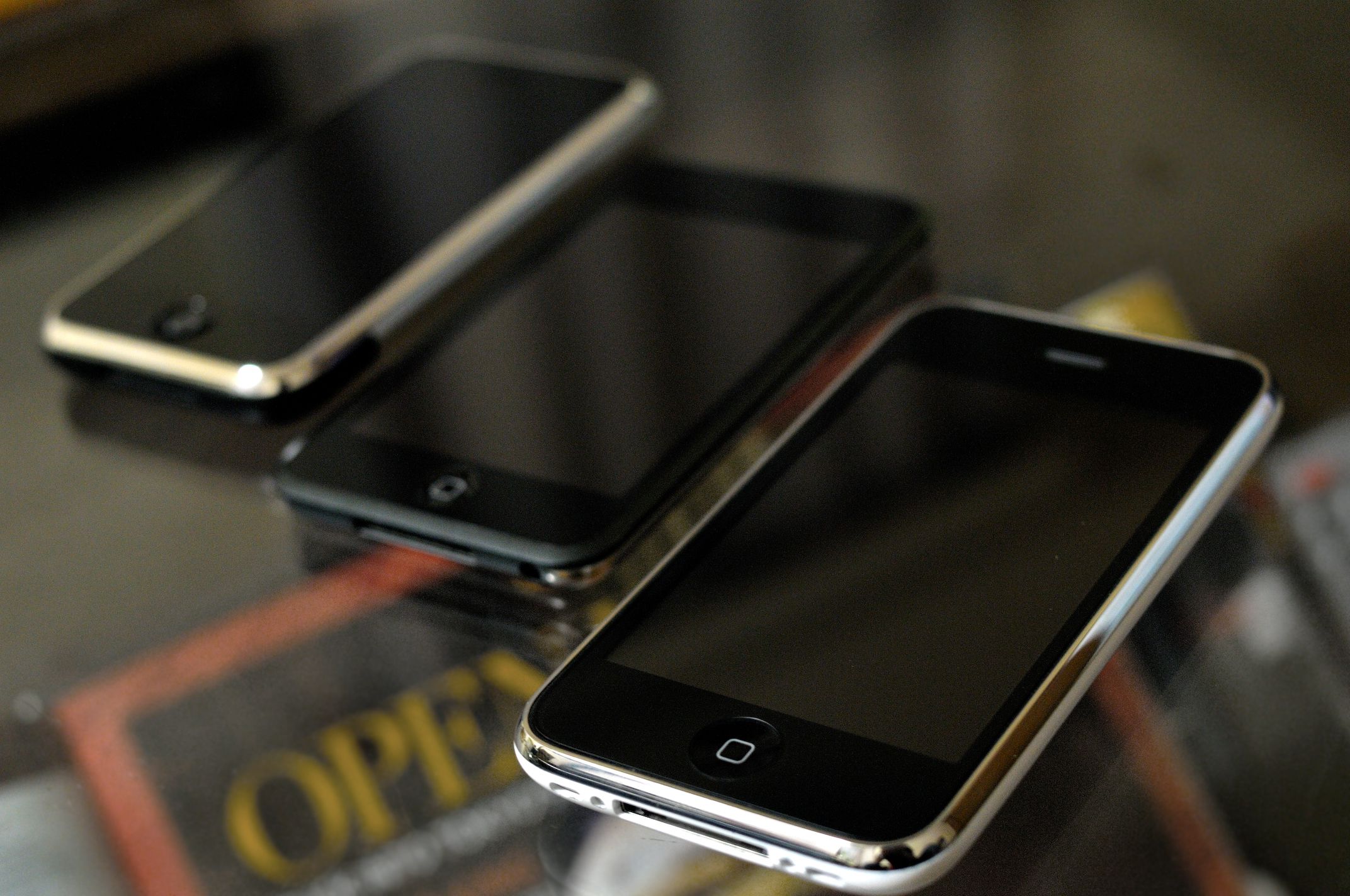 The Original iPhone, iPod Touch, and iPhone 3G on a glass table with magazines underneath