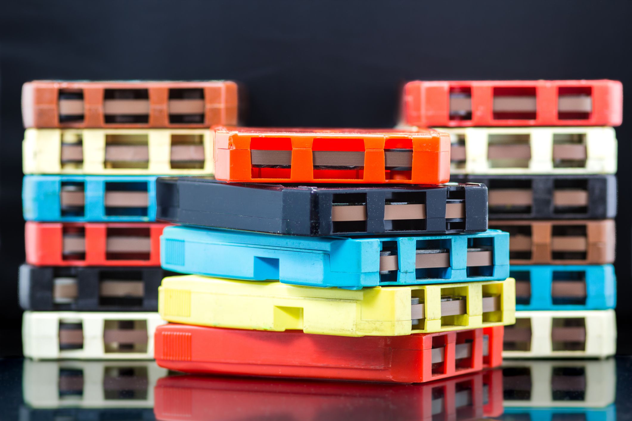 Three stacks of 8-track audio tapes in different colors, with focus on middle stack, with reflective black background