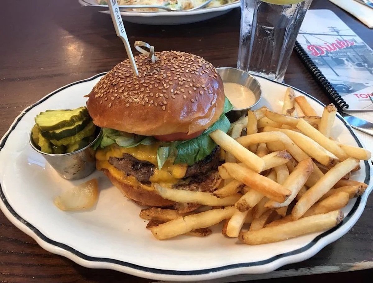 A cheeseburger with lettuce, tomato, and a sesame seed bun is served on a plate with a side of French fries, pickles, a small cup of sauce, and a glass of water in the background.