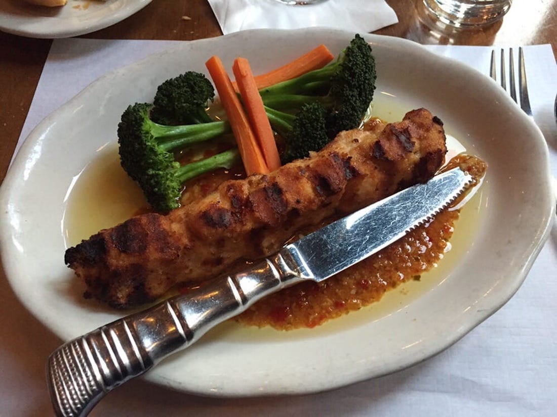 A grilled kebab skewer placed on a white plate with a knife, served with steamed broccoli and carrot sticks, and a yellow-orange sauce underneath.