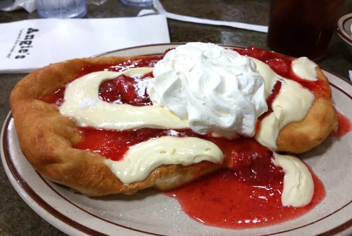 A plate with a large piece of fried dough topped with strawberry sauce, cream, and a generous swirl of whipped cream. A napkin, water glasses, and a drink are in the background.
