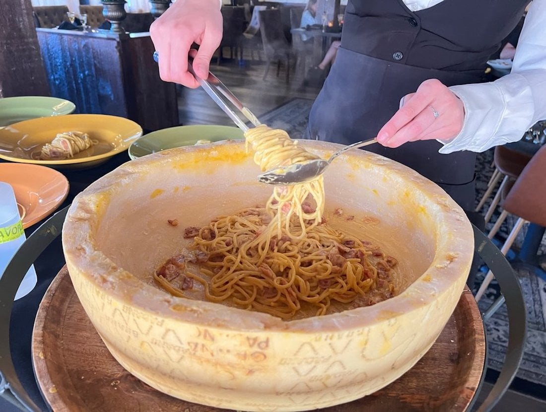 A server twirls spaghetti in a large wheel of cheese using tongs and a spoon, preparing the pasta at a restaurant table. Plates of pasta are visible in the background.