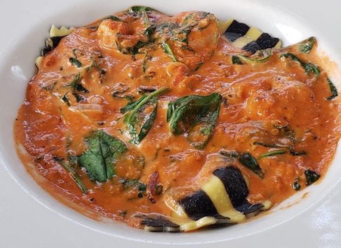 A bowl of striped black and yellow ravioli covered in a creamy tomato sauce with visible spinach leaves and herbs.