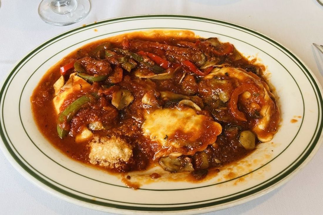 A plate of ravioli topped with tomato sauce, sliced zucchini, mushrooms, and bell peppers, served on a white plate with a green border, on a white tablecloth.